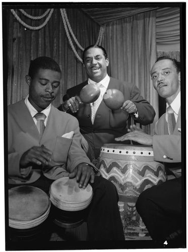 A black-and-white photograph of two musicians playing drums with their hands and one playing the maracas
